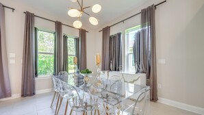 A dining area with a glass table and chairs, large windows with curtains, and a chandelier.