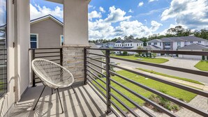 A balcony with a white chair, a stone wall, and a view of a residential area with houses and a clear sky.