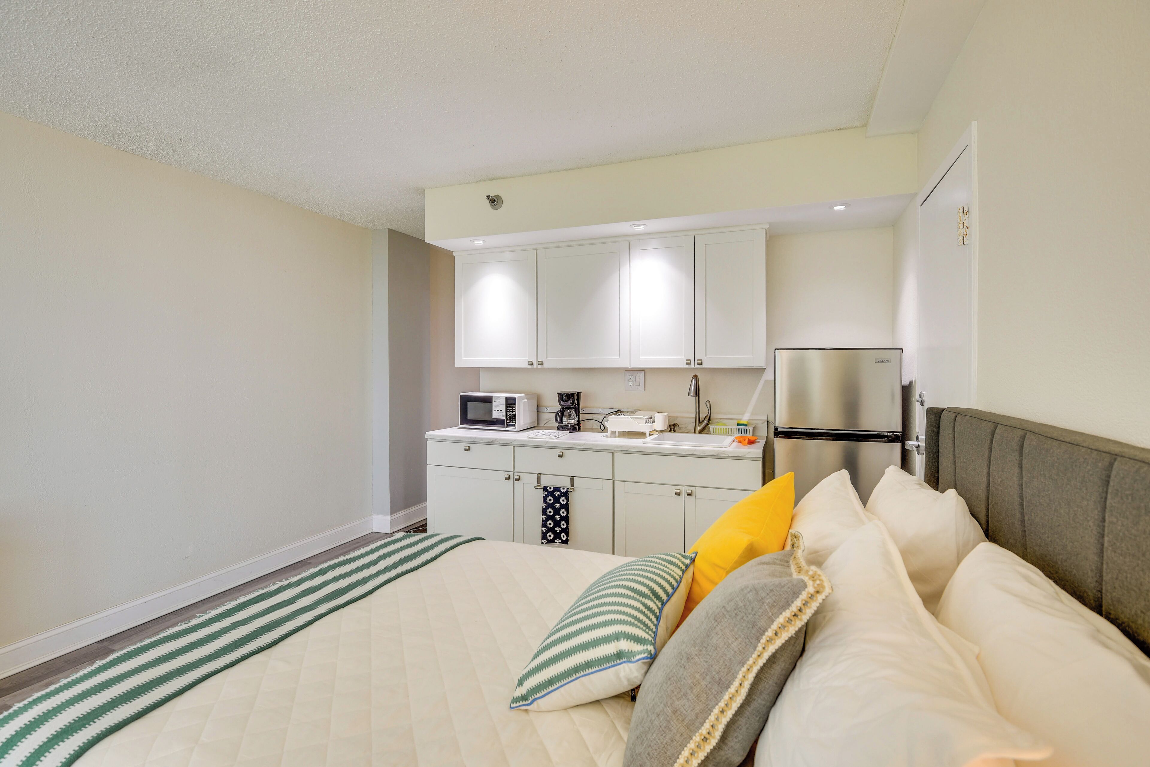 A bedroom with a bed, pillows, a bedside table, and a small kitchenette with a microwave, coffee maker, and refrigerator.