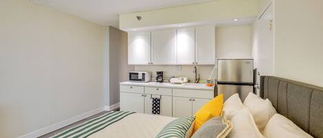 A bedroom with a bed, pillows, a bedside table, and a small kitchenette with a microwave, coffee maker, and refrigerator.