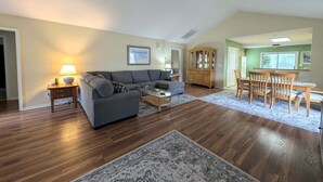 Una sala de estar con un sofá seccional gris, una mesa de centro, una mesa de comedor con sillas, un mueble de madera y un tapiz estampado.