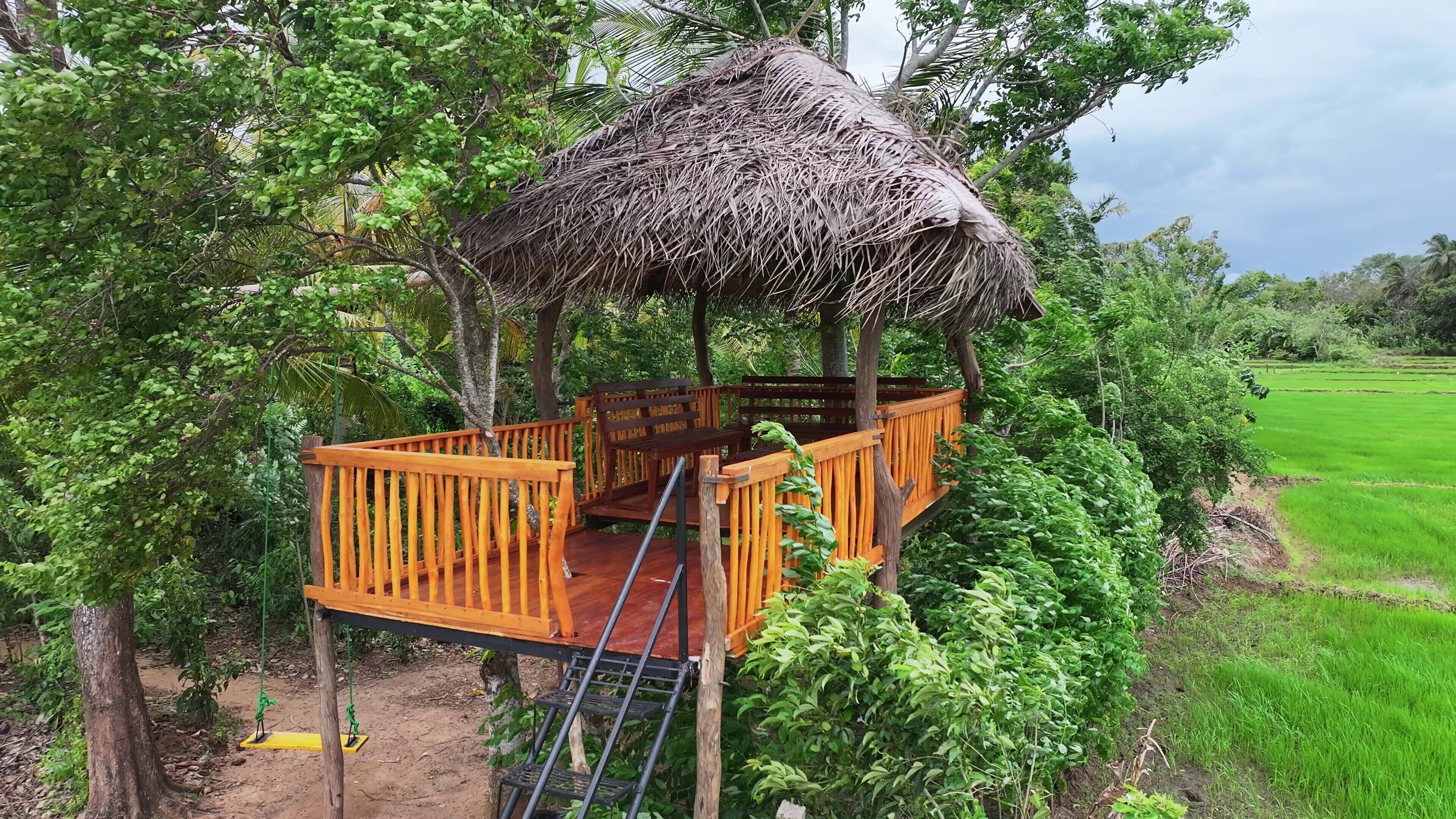 Una terraza de madera con estructura de techo de paja, rodeada de exuberante vegetación y un campo verde al fondo.
