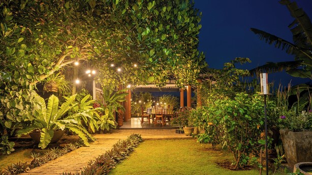 An outdoor dining area with tables and chairs under a canopy of trees, surrounded by lush greenery and a paved walkway. - Mailagama Cinnamon Residence (Sella Katharagama)
