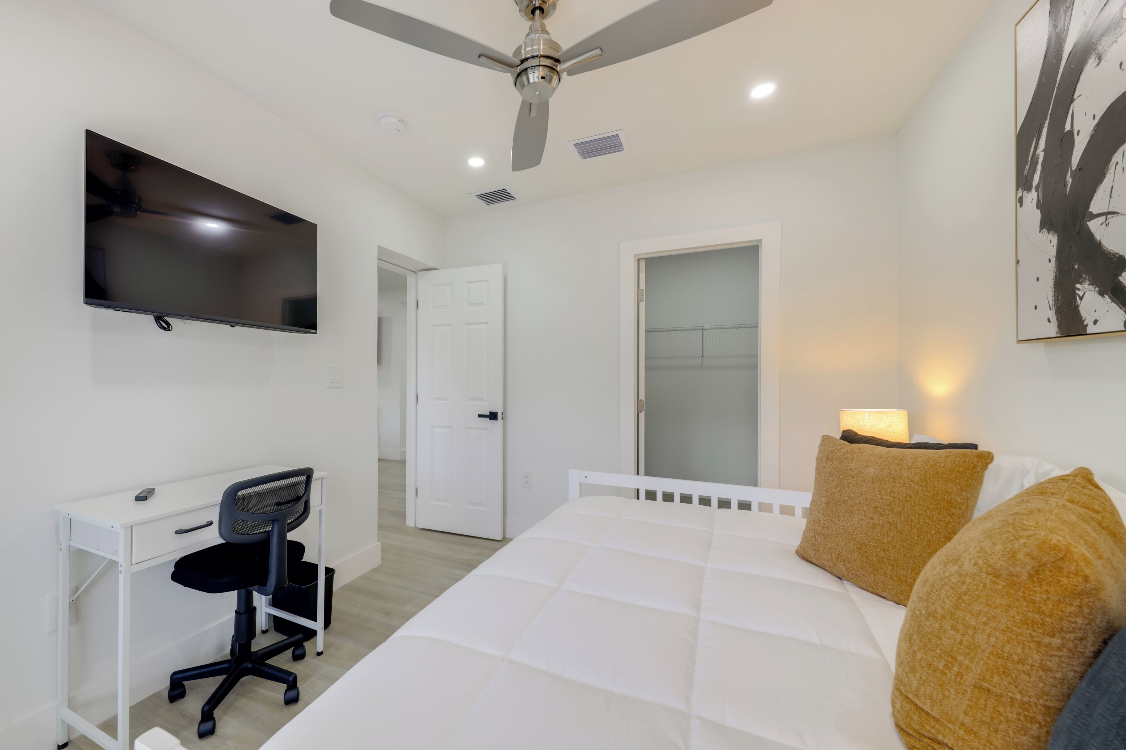 A bedroom with a bed, a desk, a chair, a ceiling fan, a TV, and a door.