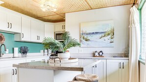 A kitchen with white cabinets, a central island with stools, a microwave, and a painting of a beach scene. - 1 Mi to Town & Rainbow Falls: Updated Hilo Getaway (Hilo)