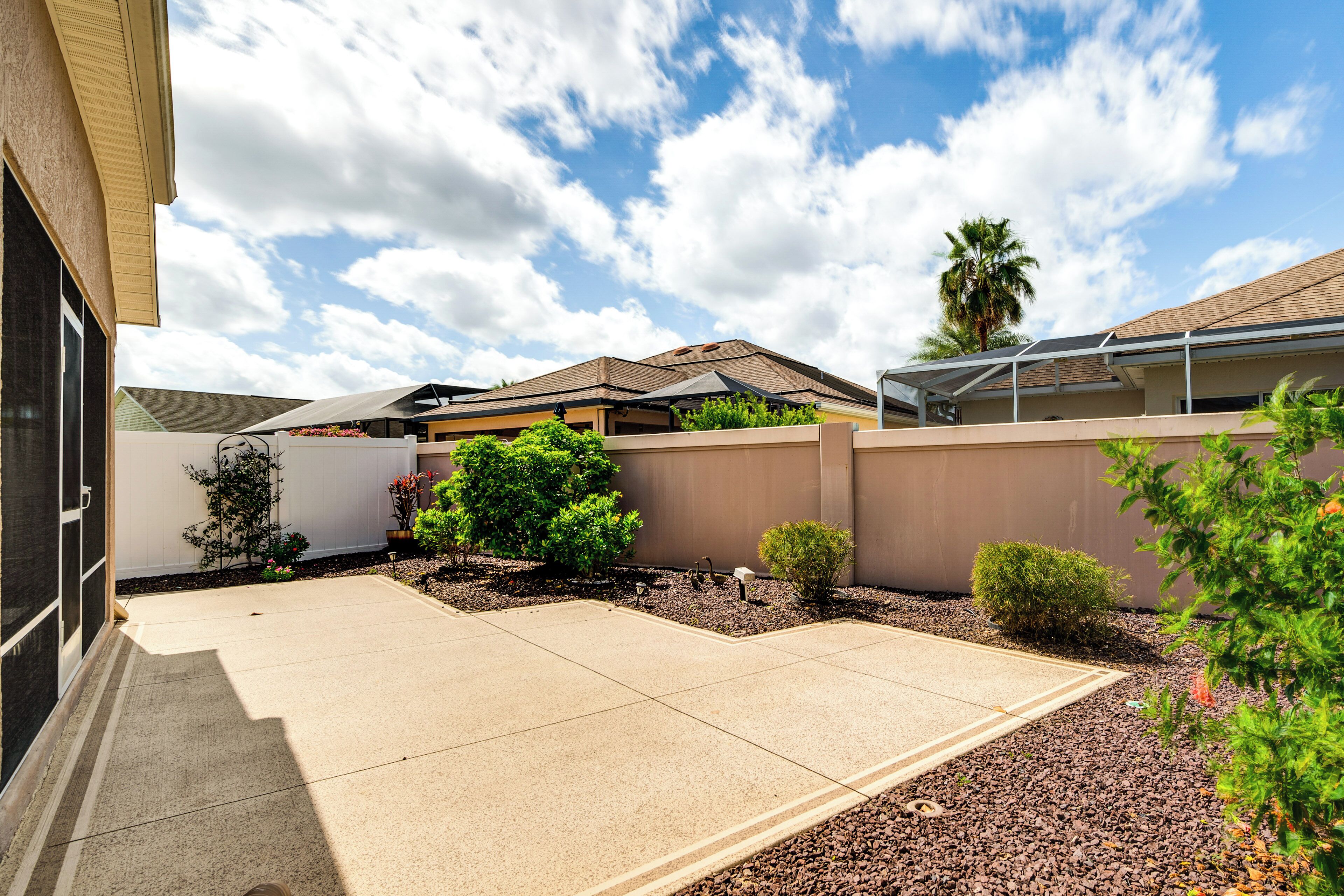 A residential area with a paved driveway, a covered patio, and a small garden with various plants and a palm tree.