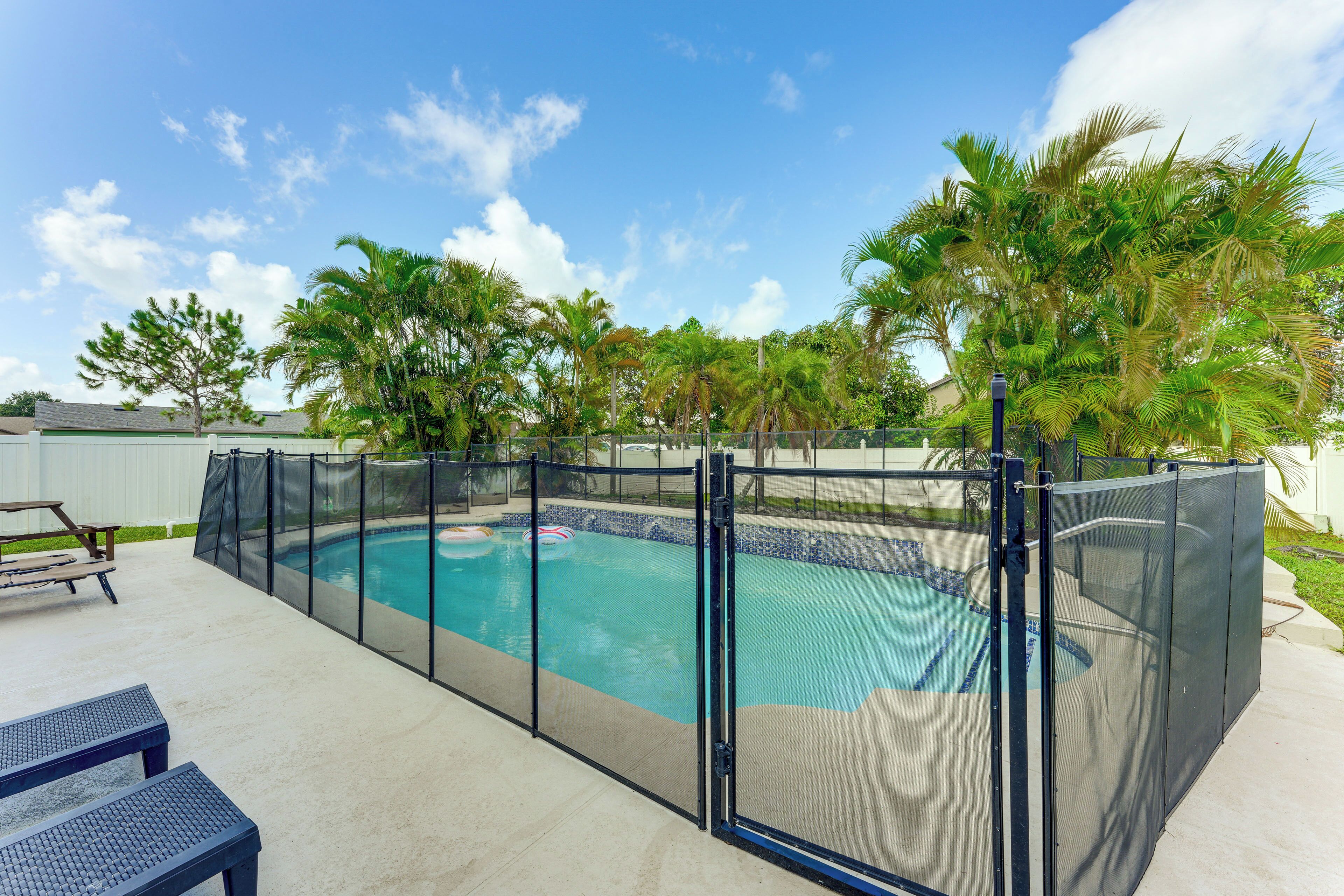 A swimming pool with a safety fence, surrounded by palm trees and a patio area with lounge chairs.
