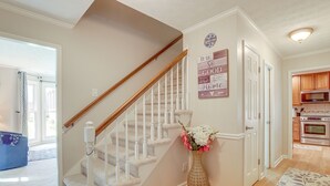 A staircase with white railings and a wooden handrail, leading to a door with a 'Welcome' sign. A vase with flowers is placed on the floor beside the staircase.