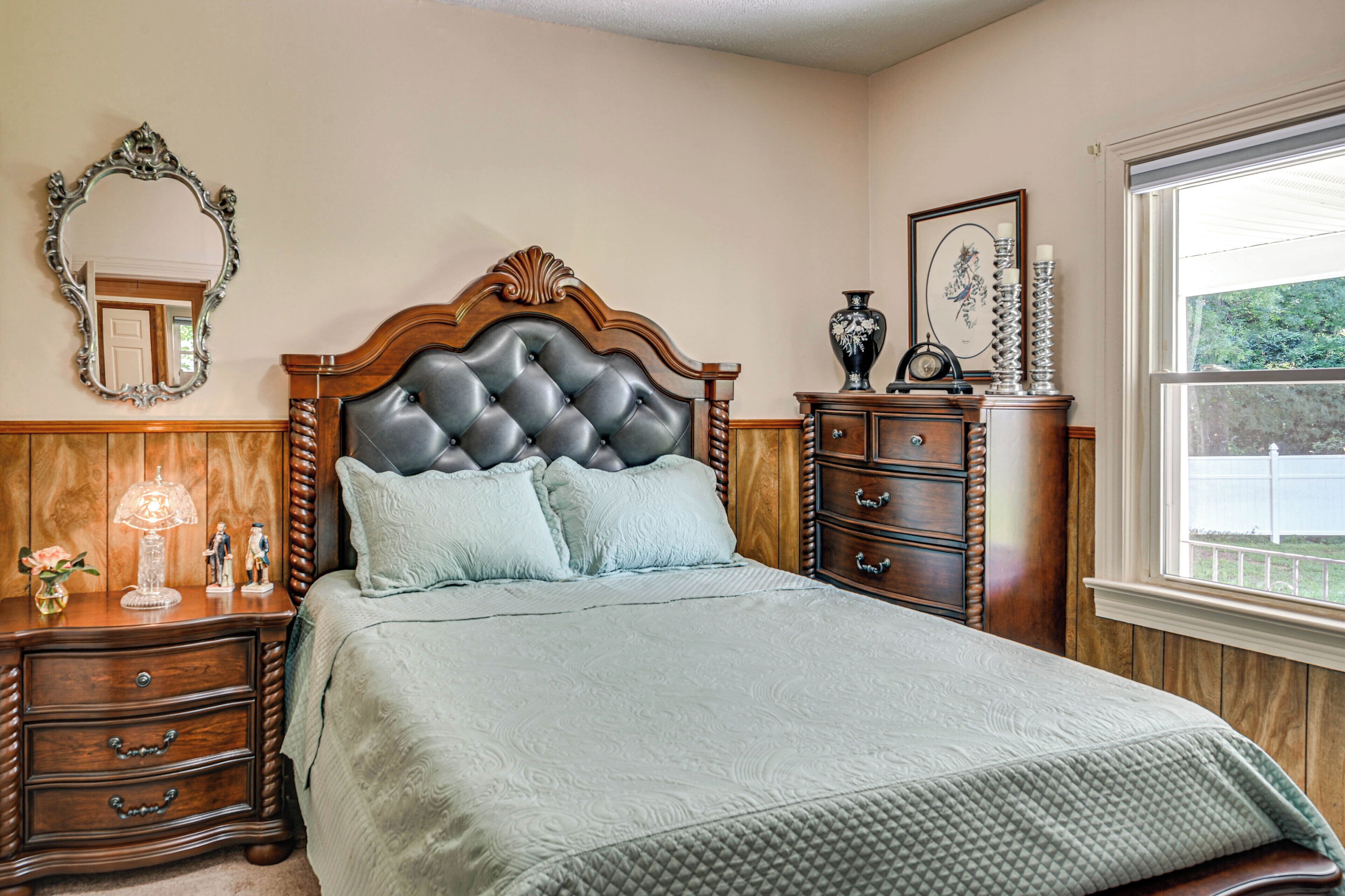 A bedroom with a tufted headboard, a large bed with a quilted cover, a nightstand with drawers, a dresser, a mirror, and a window with a view of greenery.