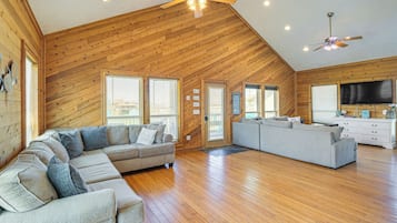 A spacious living room with a wooden ceiling, a large sectional sofa, a smaller sofa, and a TV on the wall.