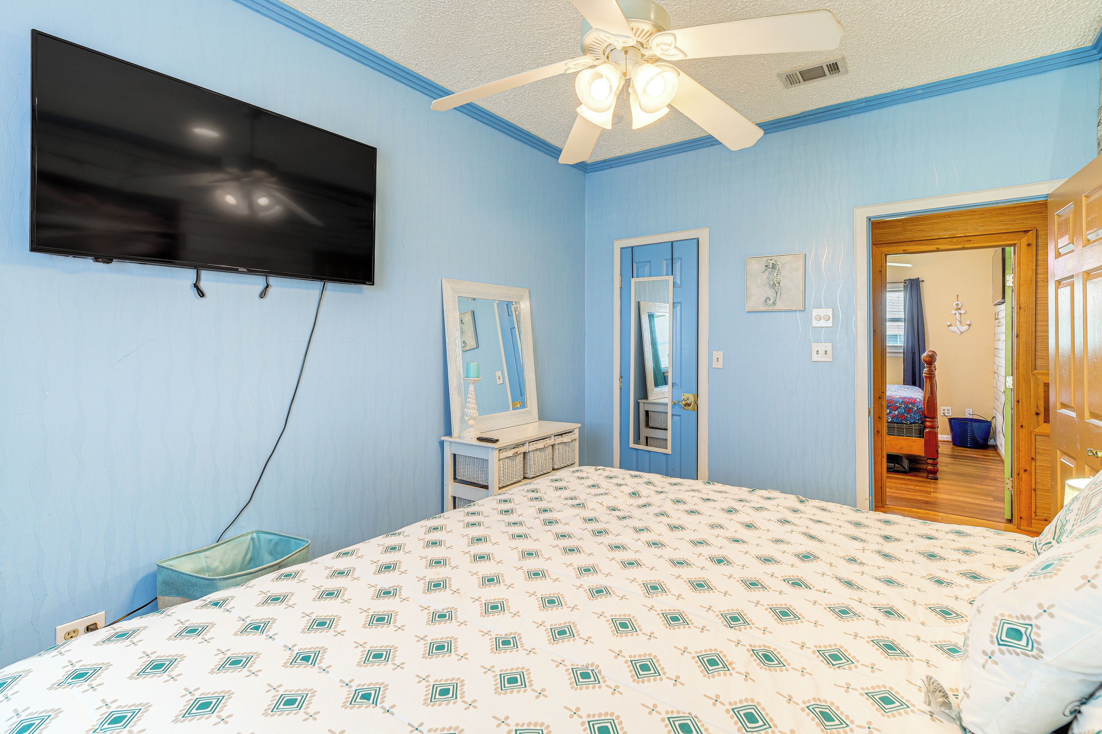 A bedroom with a bed, a television mounted on the wall, a ceiling fan, and a door leading to another room.