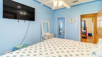 A bedroom with a bed, a television mounted on the wall, a ceiling fan, and a door leading to another room.