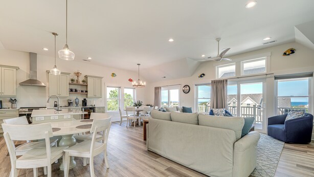 A spacious living area with a large sofa, dining table, and chairs. There is a kitchen area with modern appliances and pendant lights. The room has large windows offering a view of the outside. - Walk to Beach: Southern Shores Retreat w/ Hot Tub (Kitty Hawk)
