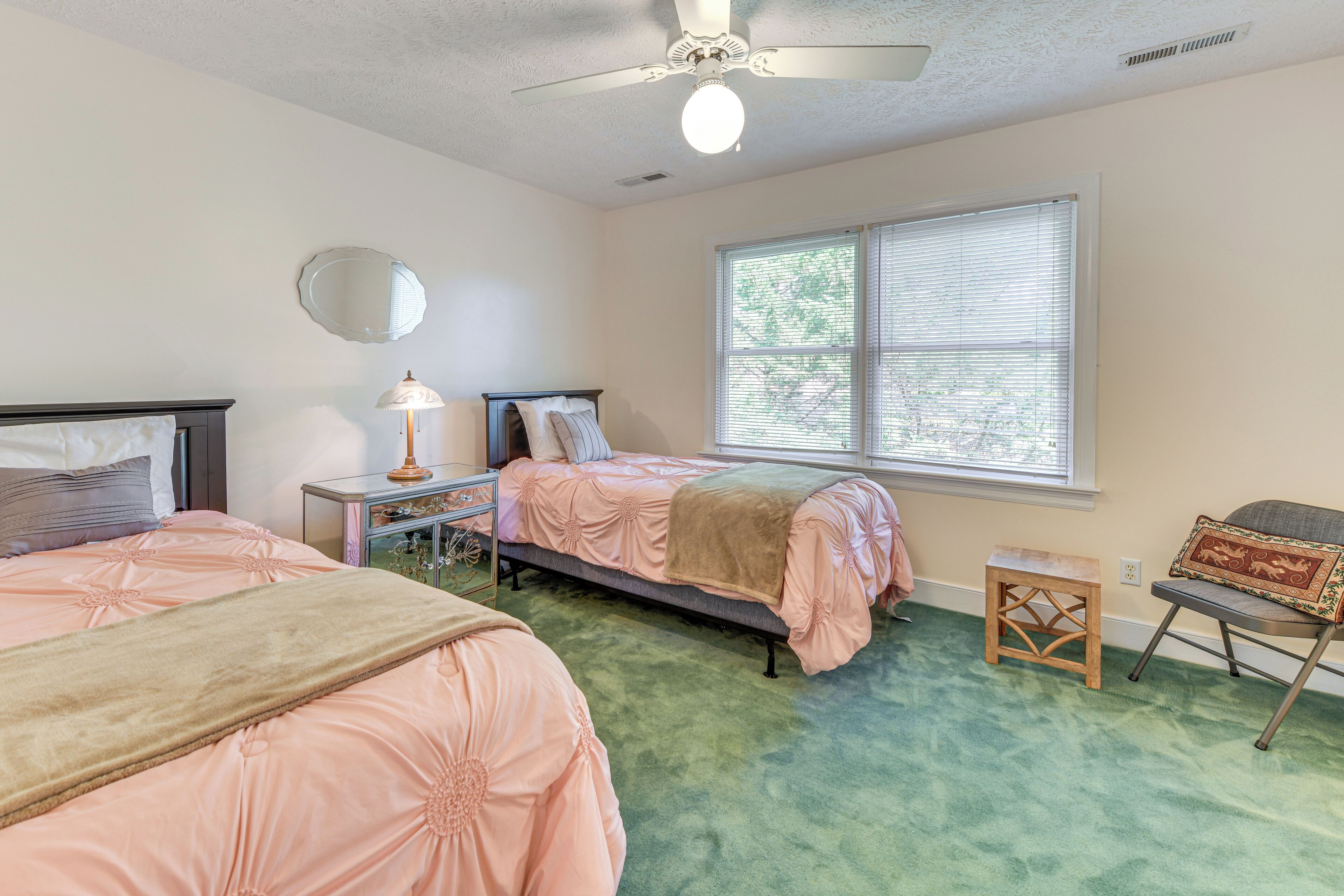 A bedroom with two beds, a ceiling fan, a window with blinds, a bedside table with a lamp, and a small stool.