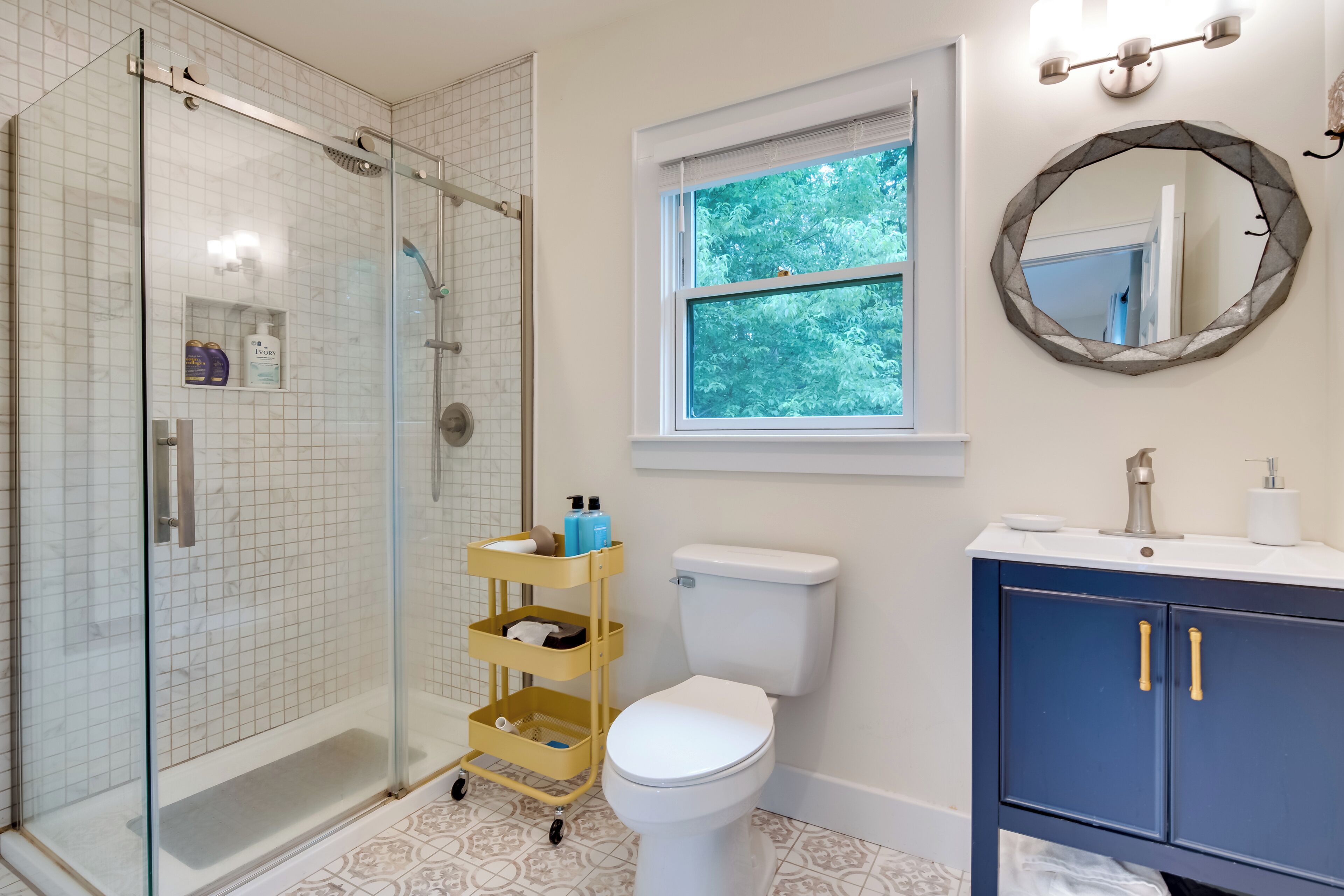 A bathroom with a glass shower enclosure, a toilet, a vanity with a sink, and a window with a view of trees.