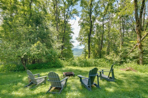 A forested area with three Adirondack chairs and a small table, surrounded by lush greenery and tall trees.