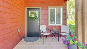 A wooden porch with a dark door, two chairs, a small table, and a potted plant with flowers. - Explore Alaskas Playground: Modern Wasilla Escape (Wasilla)