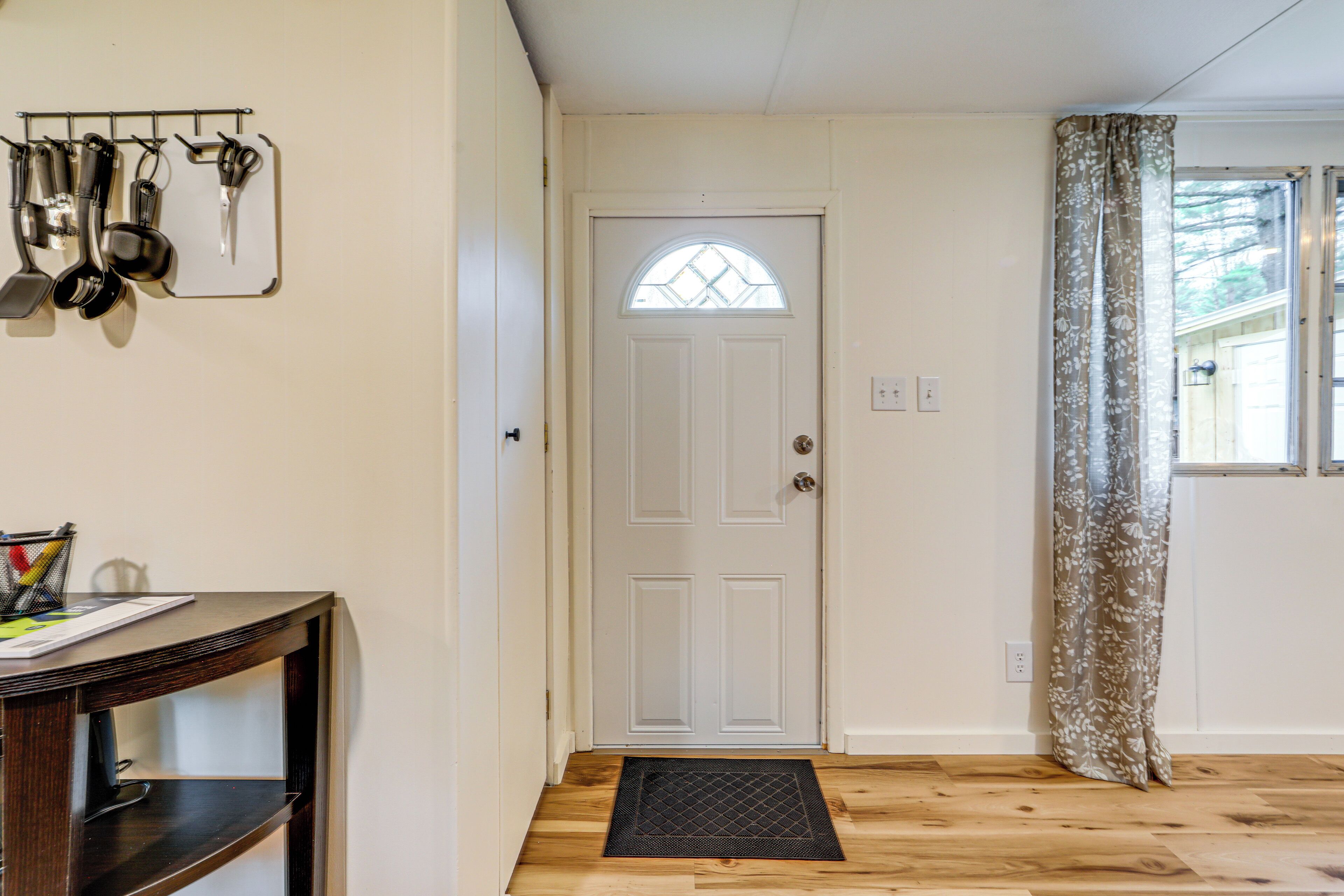 A hallway with a wooden floor, a white door with a window, and a metal rack with kitchen utensils.