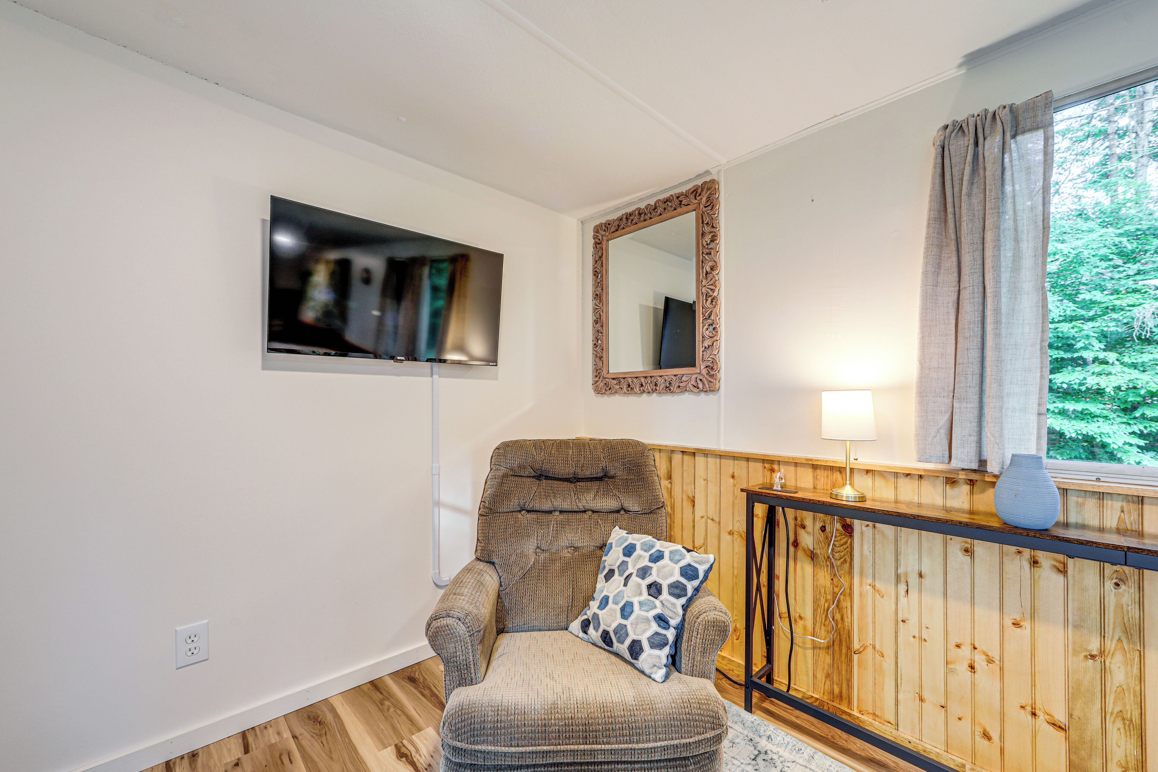 Ein Zimmer mit Holzvertäfelung, einem an der Wand montierten Fernseher, einem Sessel mit gemustertem Kissen und einem fenster mit Vorhängen.