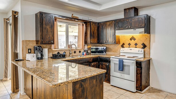 Una cocina con armarios de madera oscura, encimera de granito, estufa blanca y una ventana con cortinas color beige.