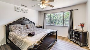 Un dormitorio con cama, ventilador de techo, ventana con cortinas y mesita de noche con un florero.