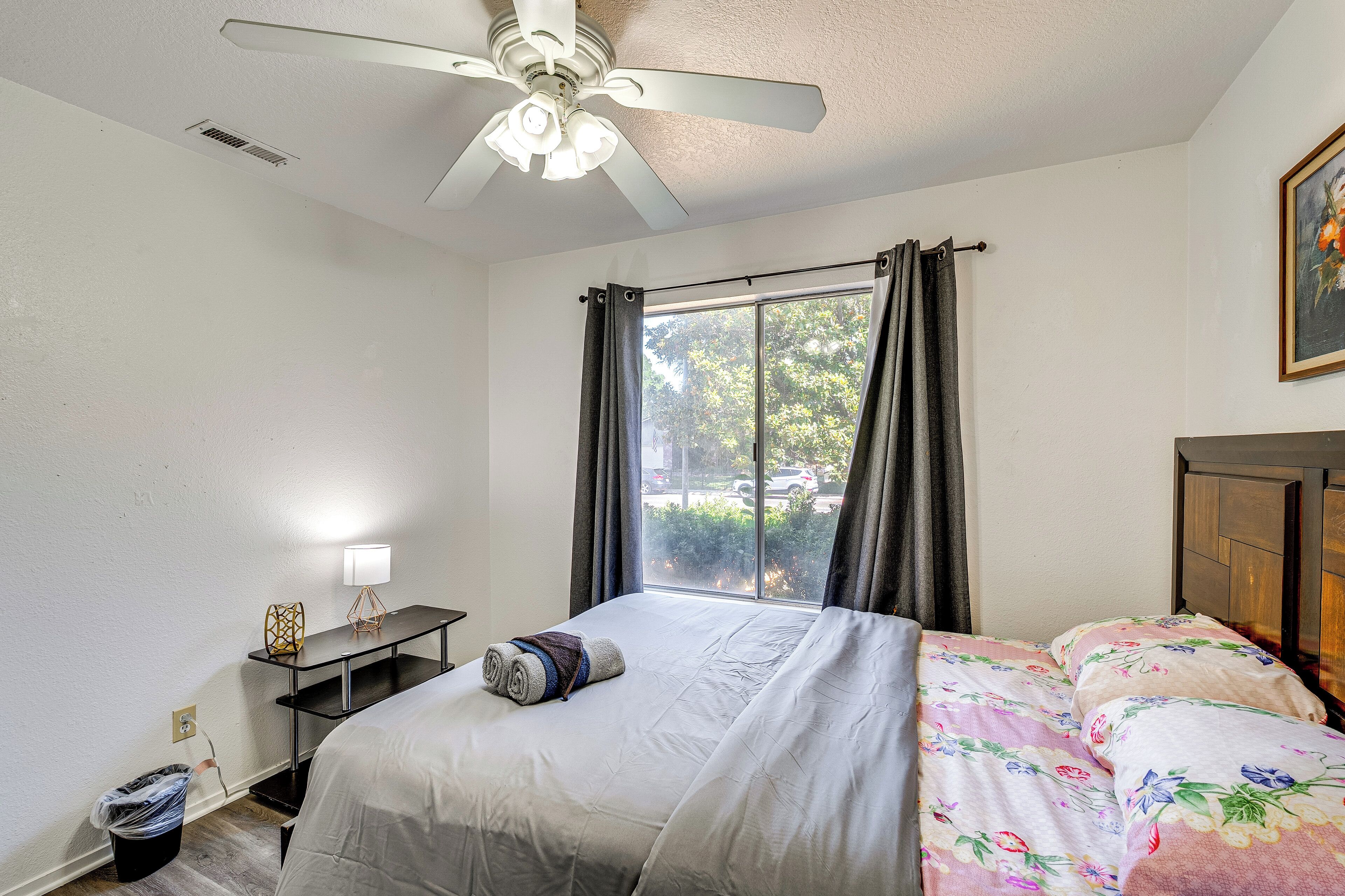 Quarto com cama, escrivaninha, ventoinha no teto, janela com cortinas e um quadro na parede.