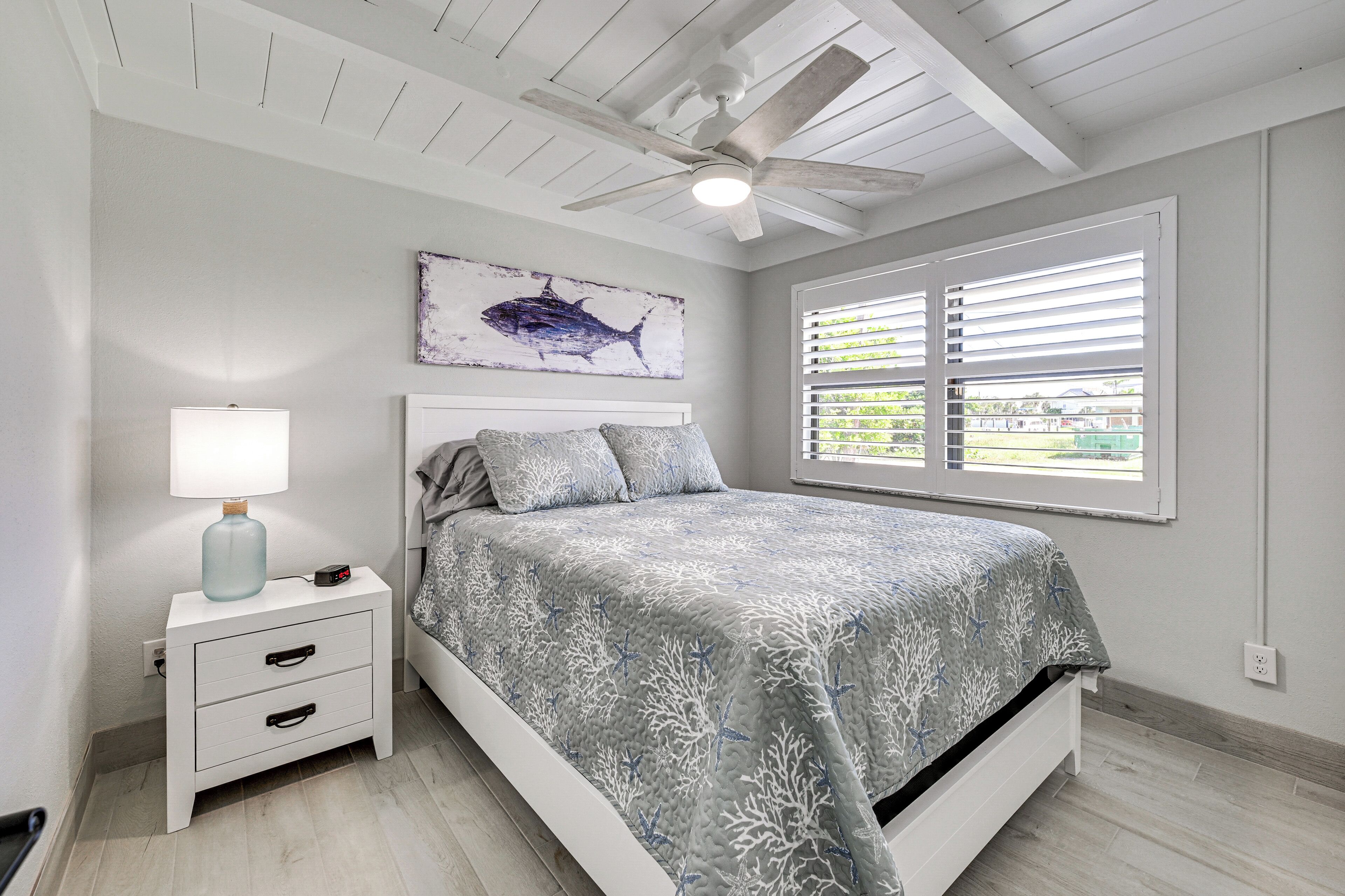 A bedroom with a bed, a nightstand with a lamp, a ceiling fan, a window with blinds, and a wall-mounted artwork of a fish.