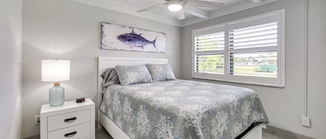 A bedroom with a bed, a nightstand with a lamp, a ceiling fan, a window with blinds, and a wall-mounted artwork of a fish.