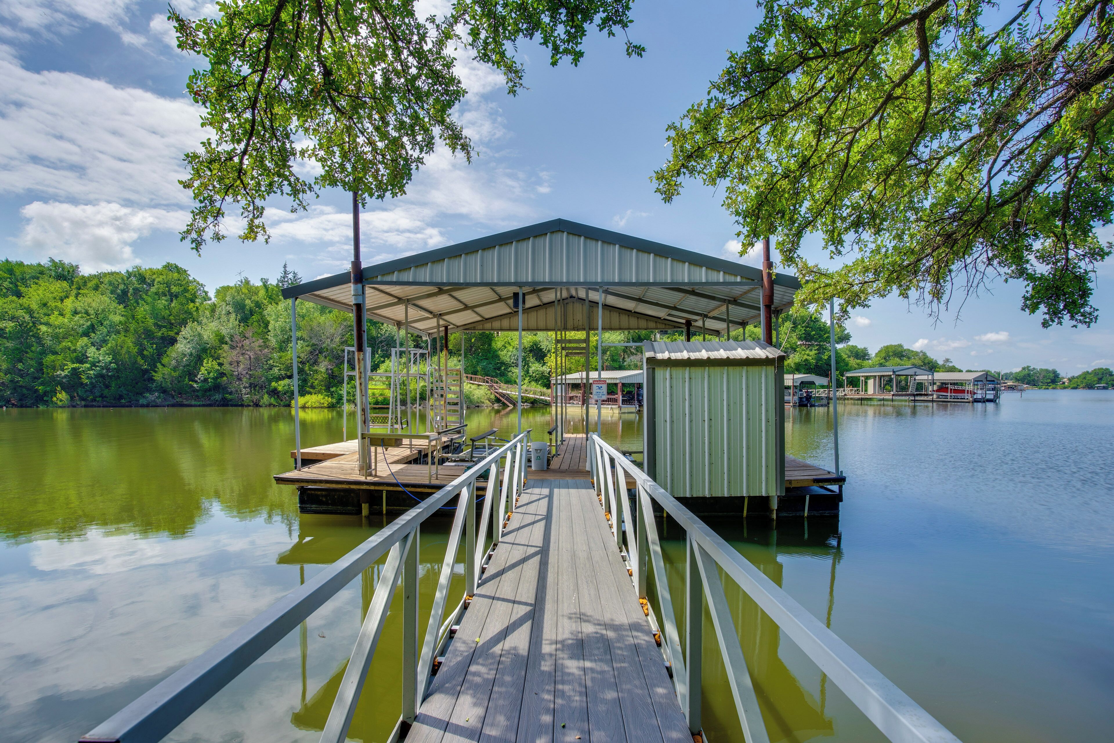 A wooden dock leading to a covered pavilion by a calm lake, surrounded by lush greenery.