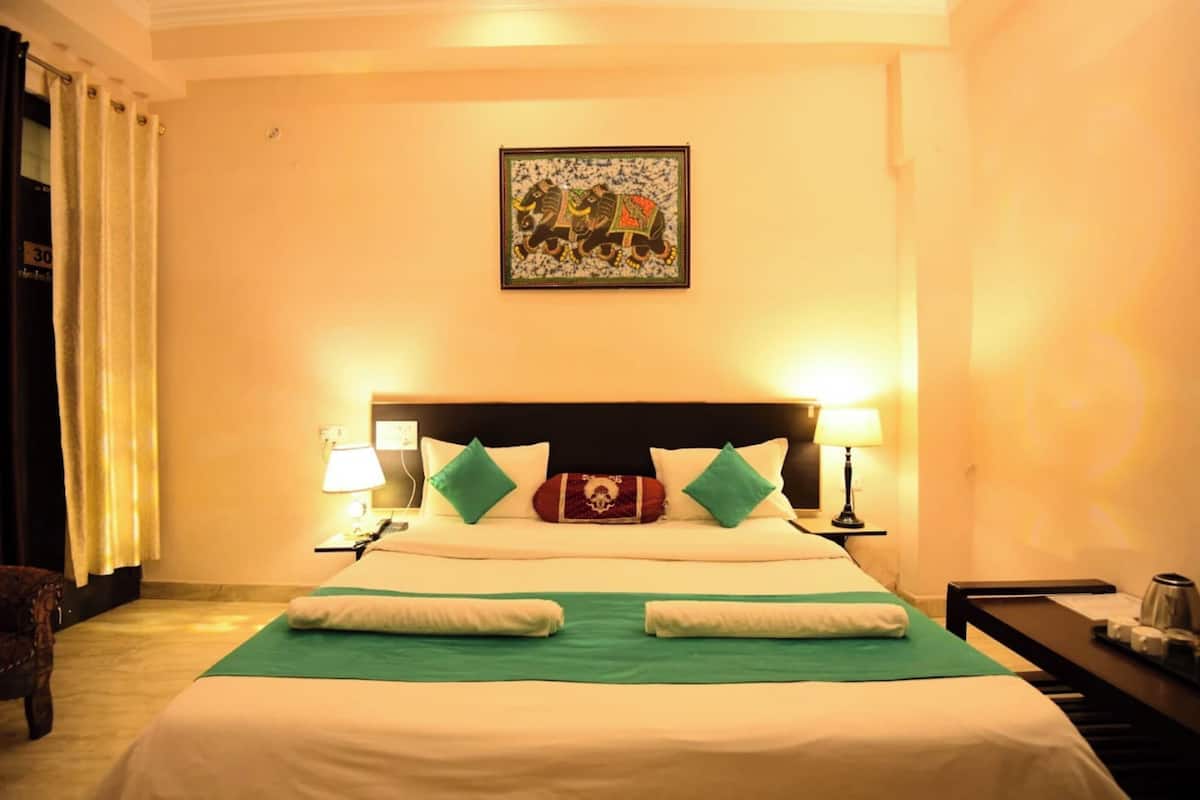 Quarto de hotel com uma cama grande, duas luminárias de cabeceira, um travesseiro decorativo, uma obra de arte emoldurada na parede e uma pequena mesa com uma chaleira.