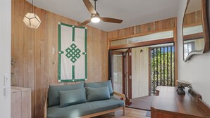 Ein Zimmer mit Holzboden, einem grünen Sofa, einem Deckenventilator und einem großen Fenster mit Blick ins Grüne.