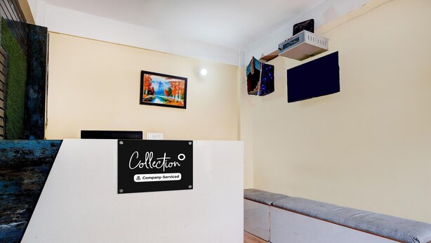 A reception area with a sign indicating it's a company-serviced collection point, a bench, a wall-mounted shelf with a small item, and a framed picture on the wall. - Collection O Last Gate Formerly RR Hotels (Guwahati)