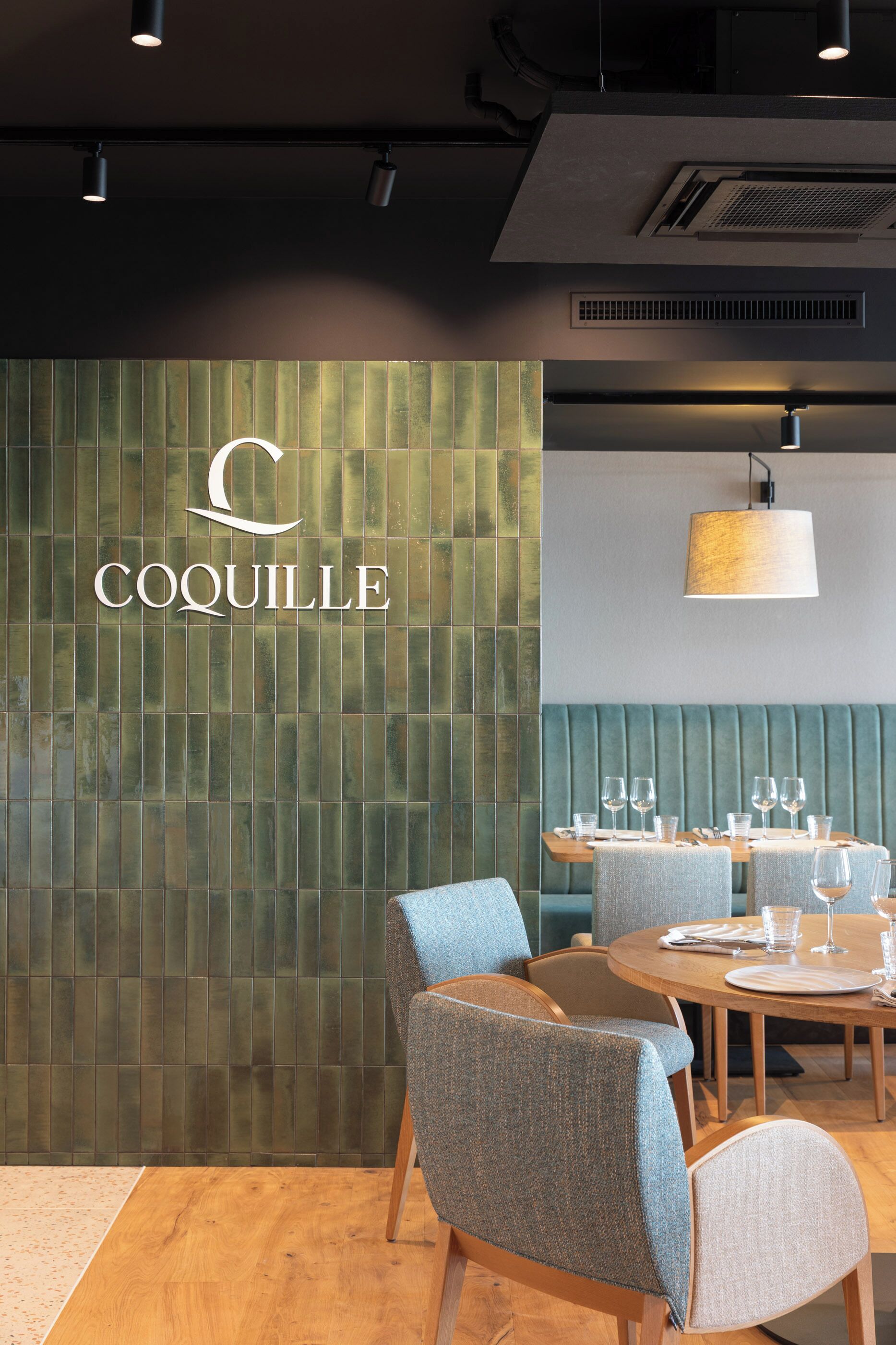 Ein Restaurantinneres mit einer grünen Wand aus Fliesen, einem Holztisch, der mit Gläsern und Besteck gedeckt ist, und einer blauen Polsterbank.