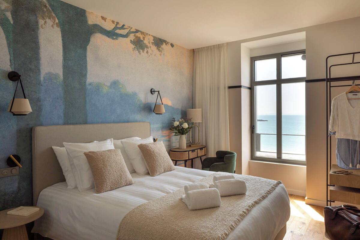 Une chambre d’hôtel avec un grand lit, une vue sur la mer, un bureau avec une lampe et une chaise.
