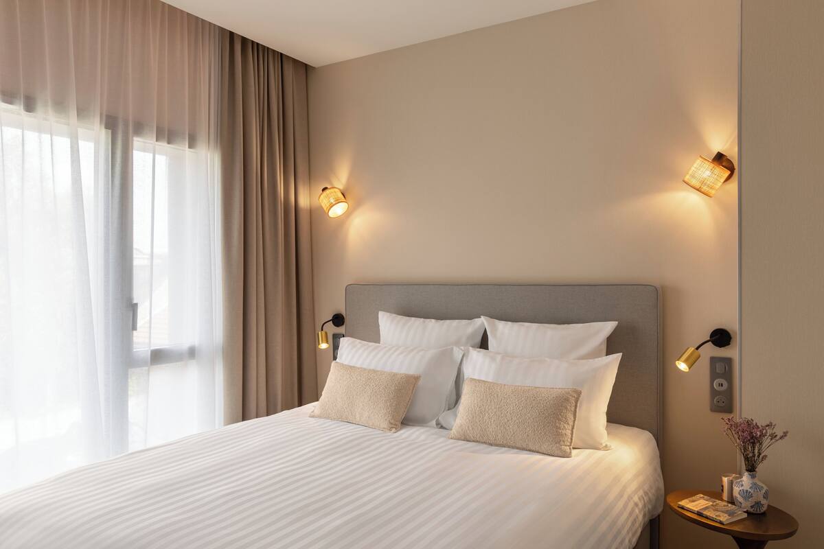Un lit bien fait, avec du linge de lit blanc et des oreillers beige, une table de chevet ornée d’un vase et d’un livre, et deux lampes fixées au mur, de part et d’autre du lit.