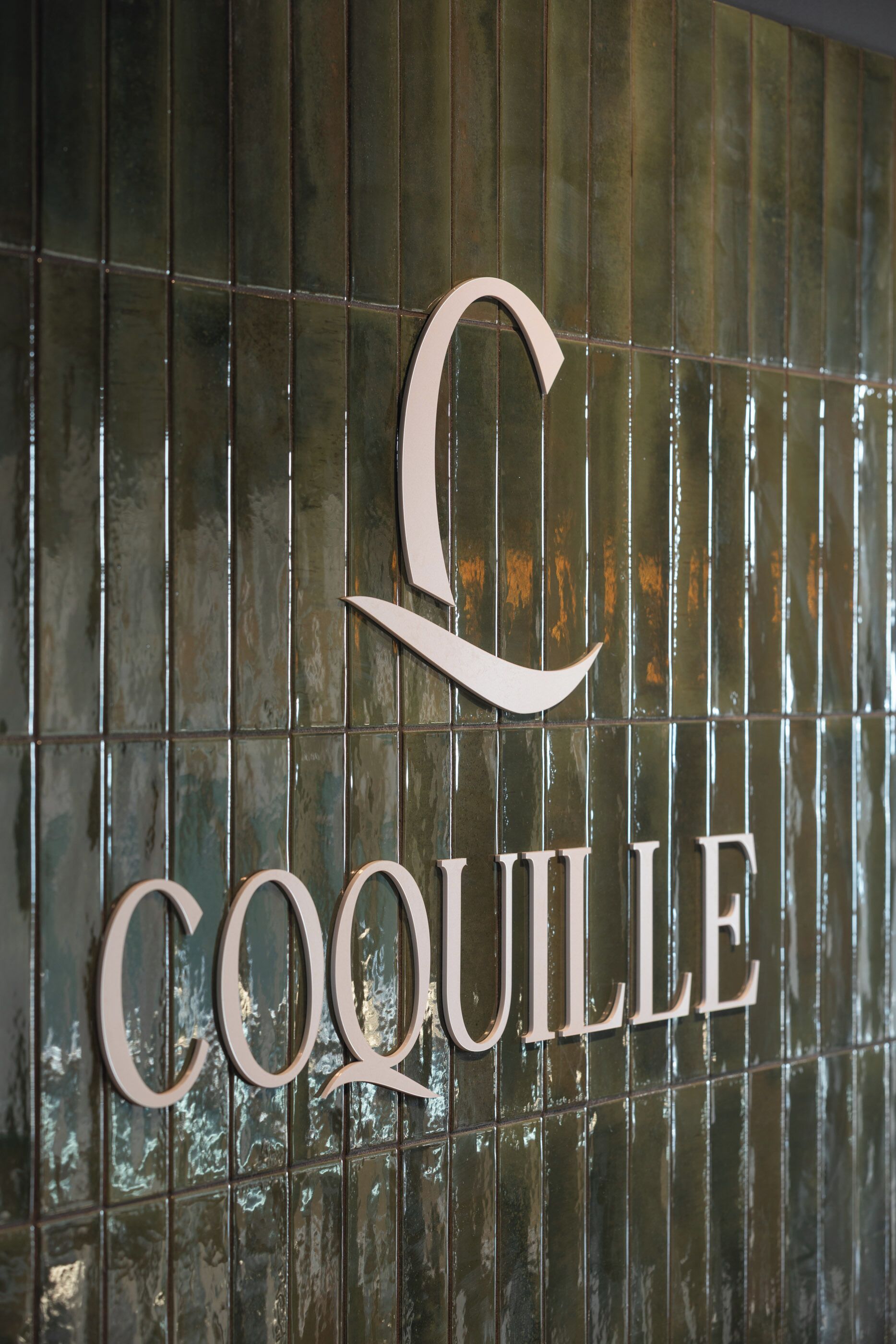 En skylt med ordet ”COQUILLE” skrivet med stora, eleganta bokstäver på en kakelbeklädd vägg.