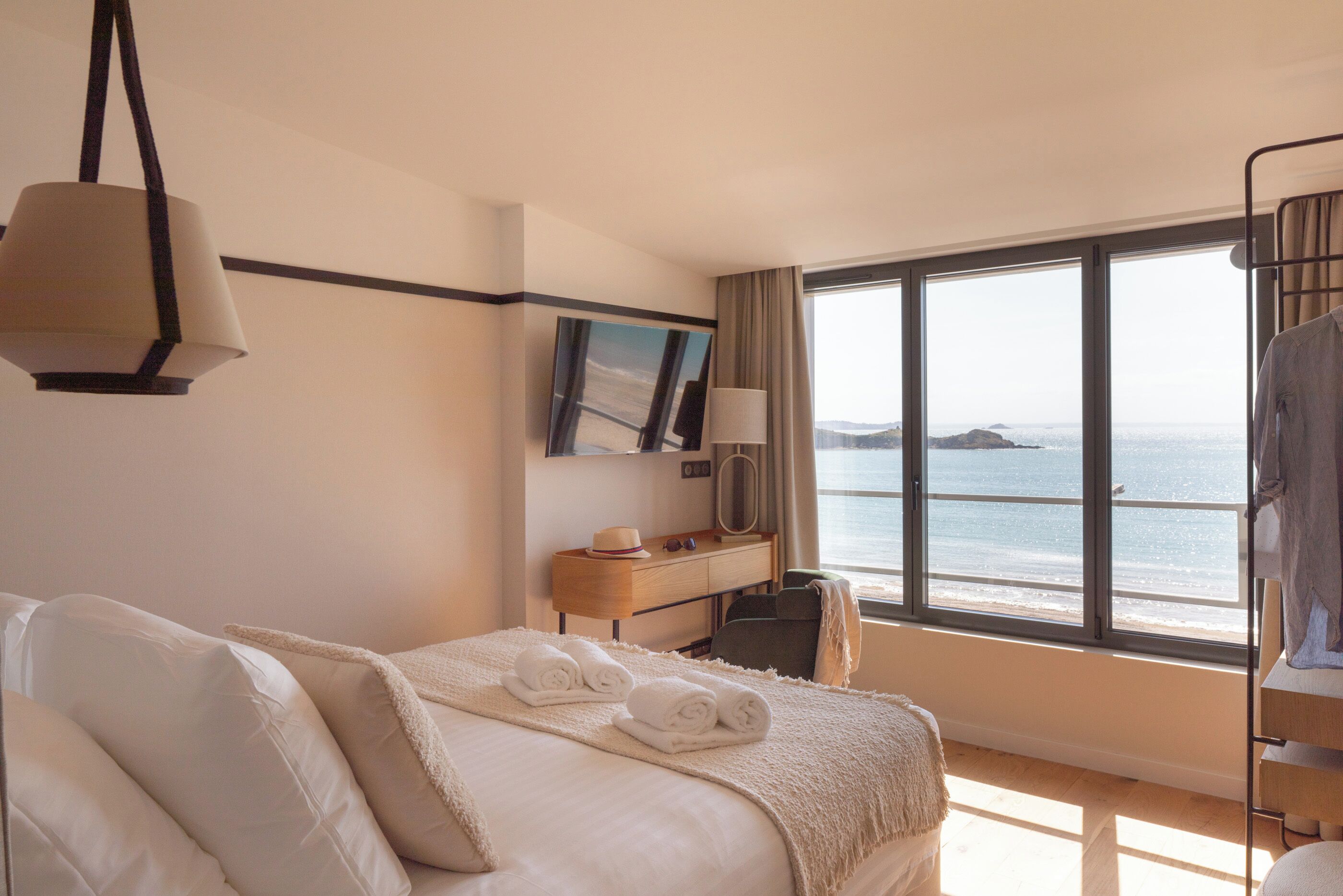 Une chambre d’hôtel comprenant un lit, un bureau, une chaise, une lampe, une fenêtre donnant sur la mer et une suspension.