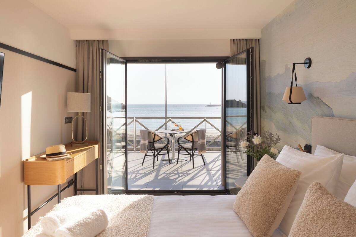 Une chambre d’hôtel avec un grand lit, un bureau avec une lampe, une vue sur la mer par une porte vitrée et un tableau au mur.