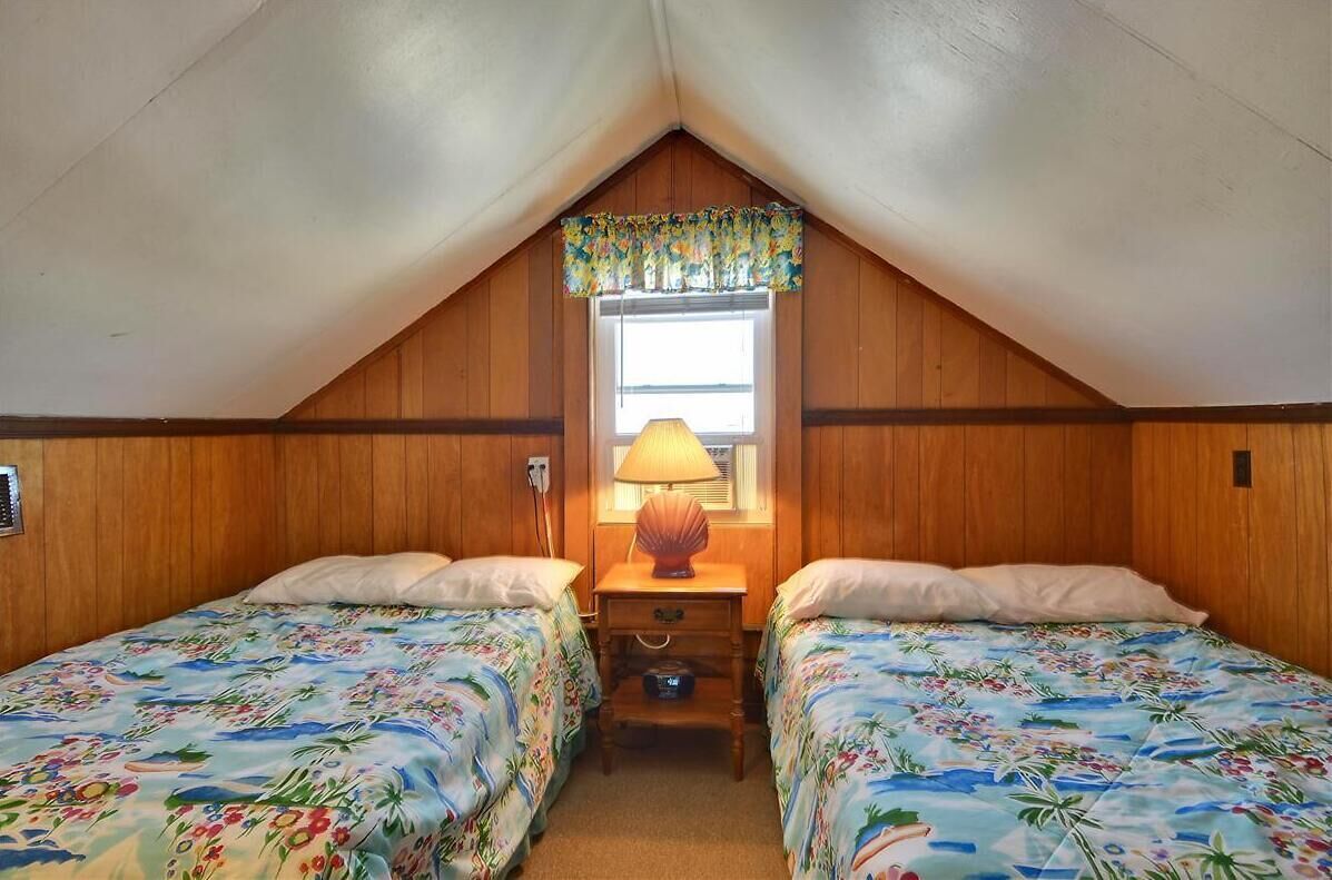 A small, cozy room with two beds, a window with a floral valance, and a lamp on a nightstand.