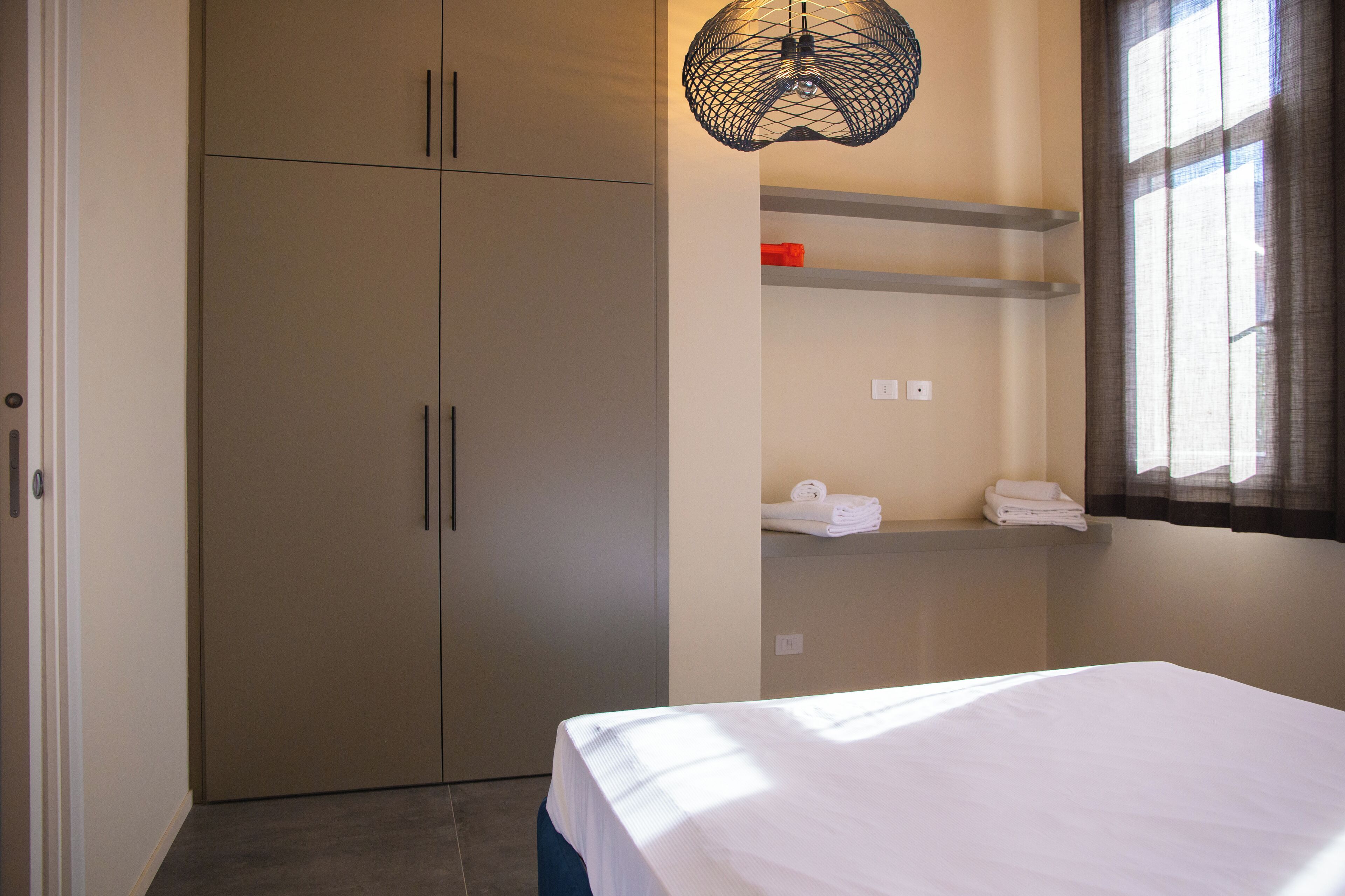 A bedroom with a bed, a wardrobe, a shelf with towels, and a hanging light fixture.