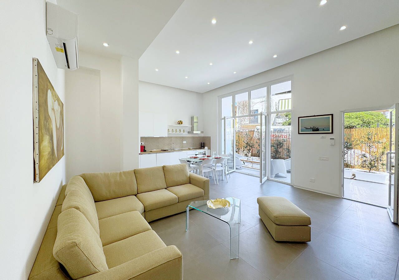 A modern living room with a beige sectional sofa, a glass coffee table, and a small ottoman. There is a kitchen area in the background with white cabinets and a dining table.