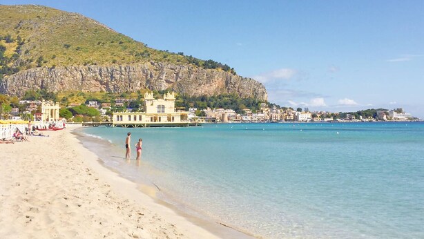 Une plage à l’eau turquoise et limpide, une rive sablonneuse et, au loin, un bâtiment à la façade jaune.