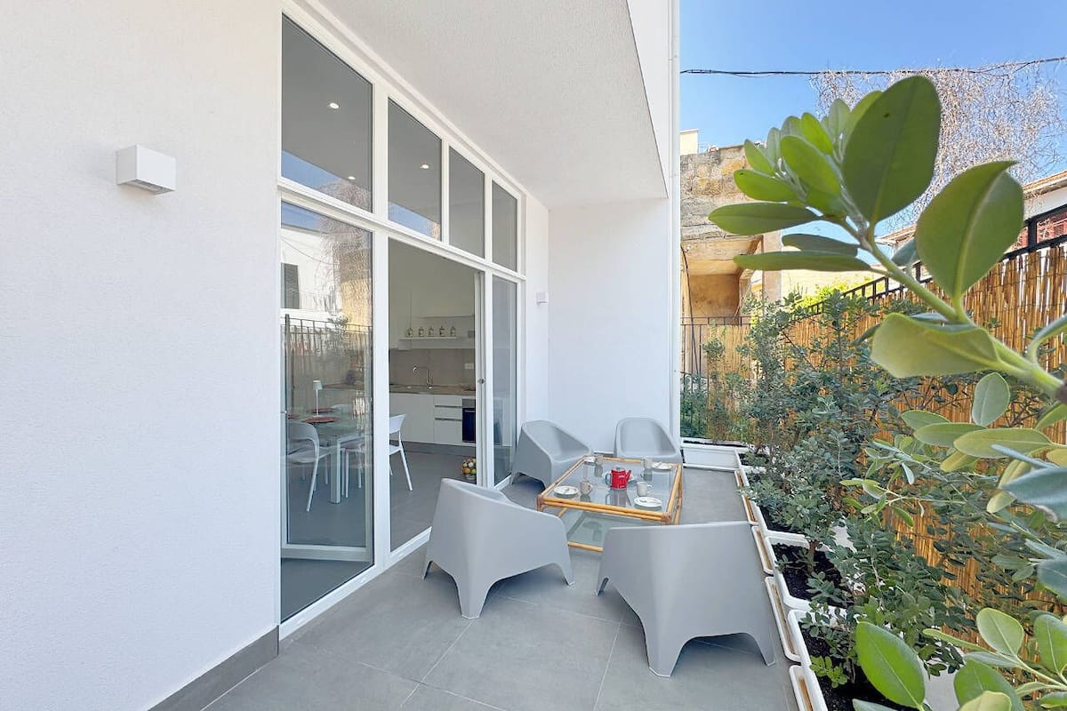 Une terrasse extérieure moderne avec une table et des chaises en verre, un petit espace jardin agrémenté de plantes et une vue sur un bâtiment en arrière-plan.