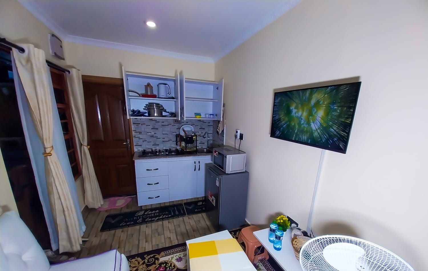 A compact kitchen with a gas stove, microwave, and a small refrigerator. There is a dining area with a table and chairs, and a wall-mounted TV.
