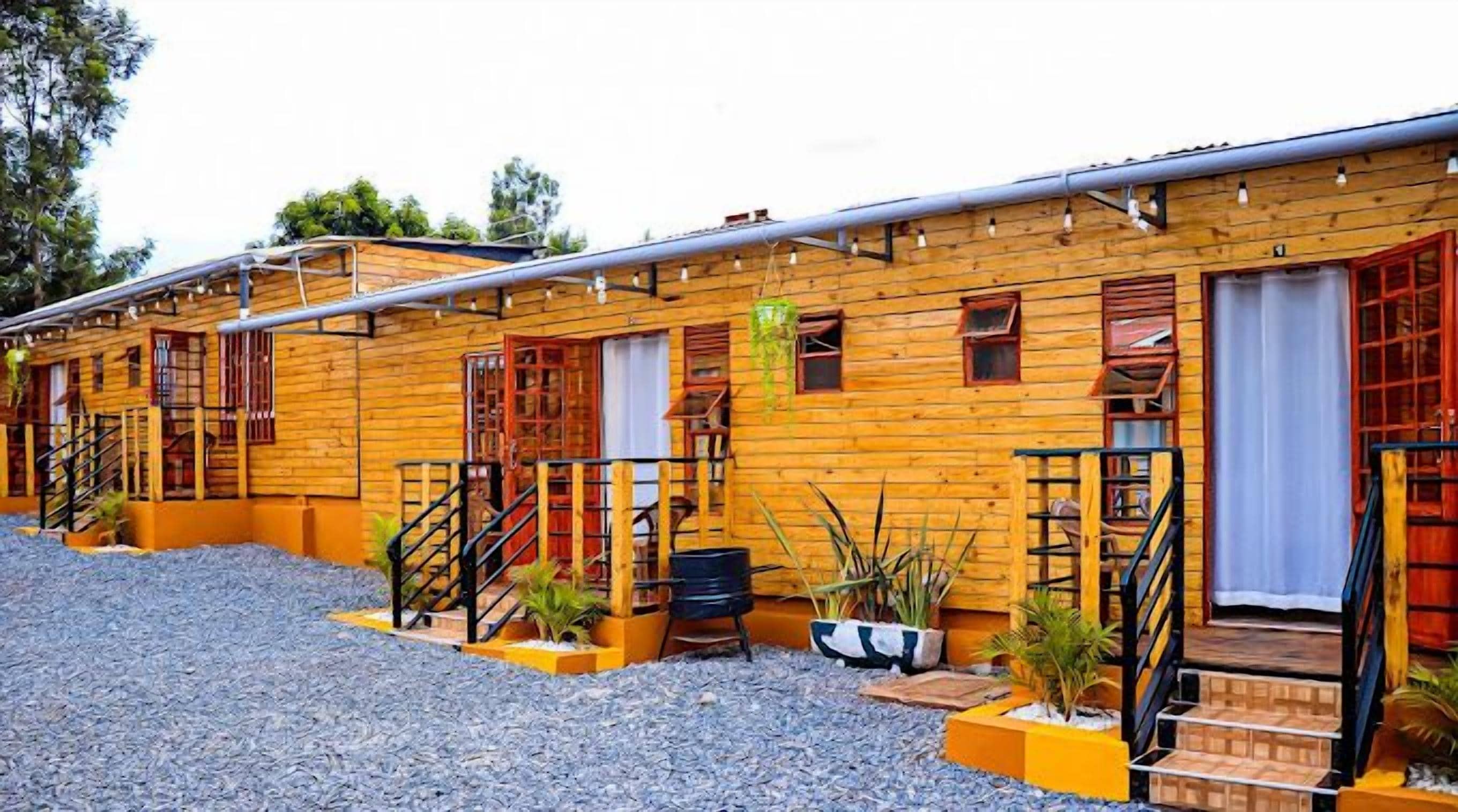 Filas de cabañas de madera con escaleras exteriores, un camino de grava y macetas con plantas.