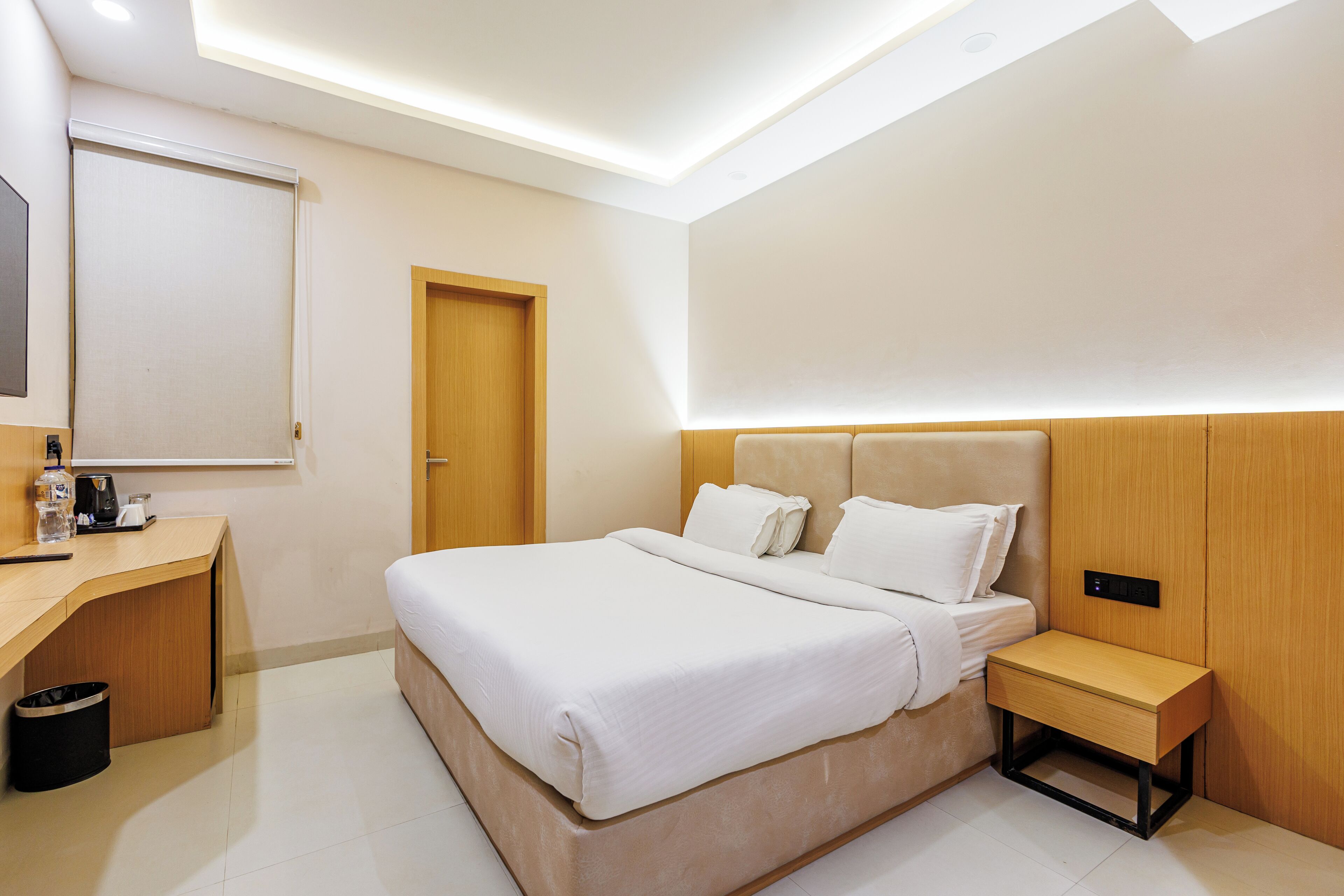 A modern hotel room with a large bed, a wooden desk, a nightstand, and a wall-mounted control panel.