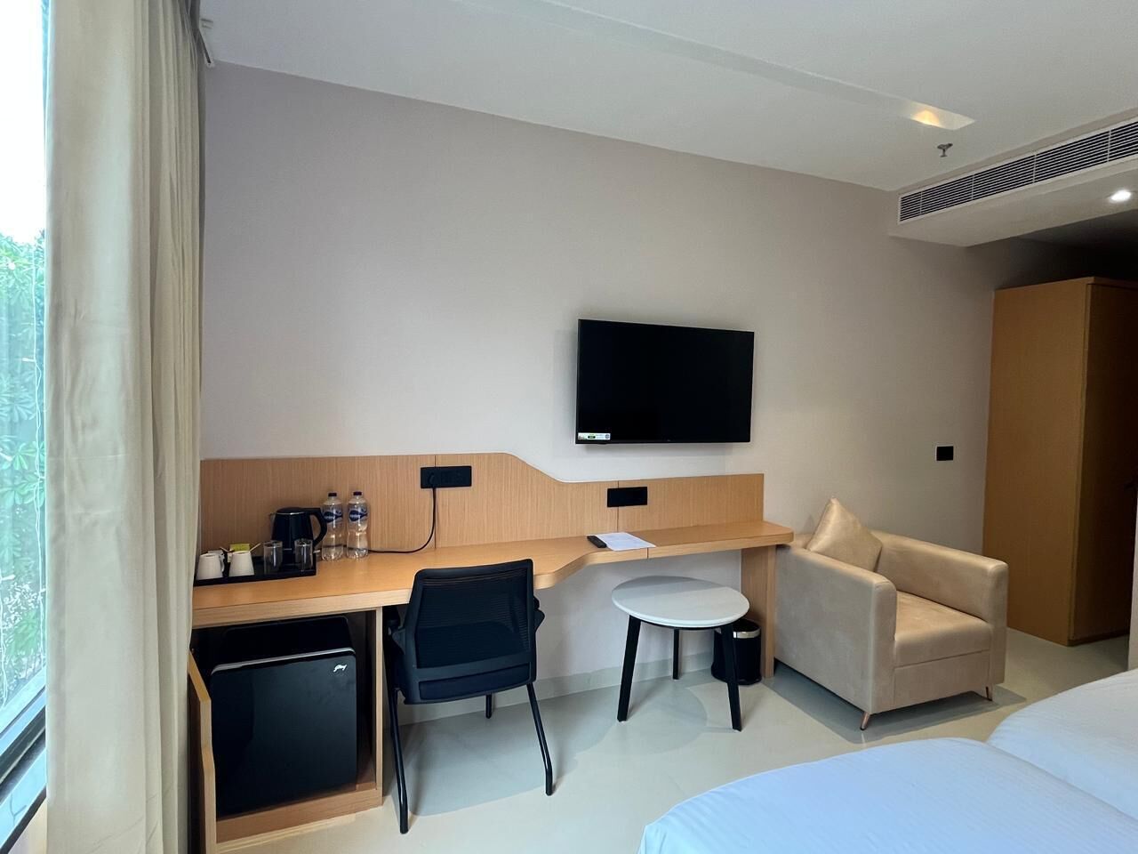 A hotel room with a bed, a desk with a chair, a small round table, a television mounted on the wall, and a wardrobe.