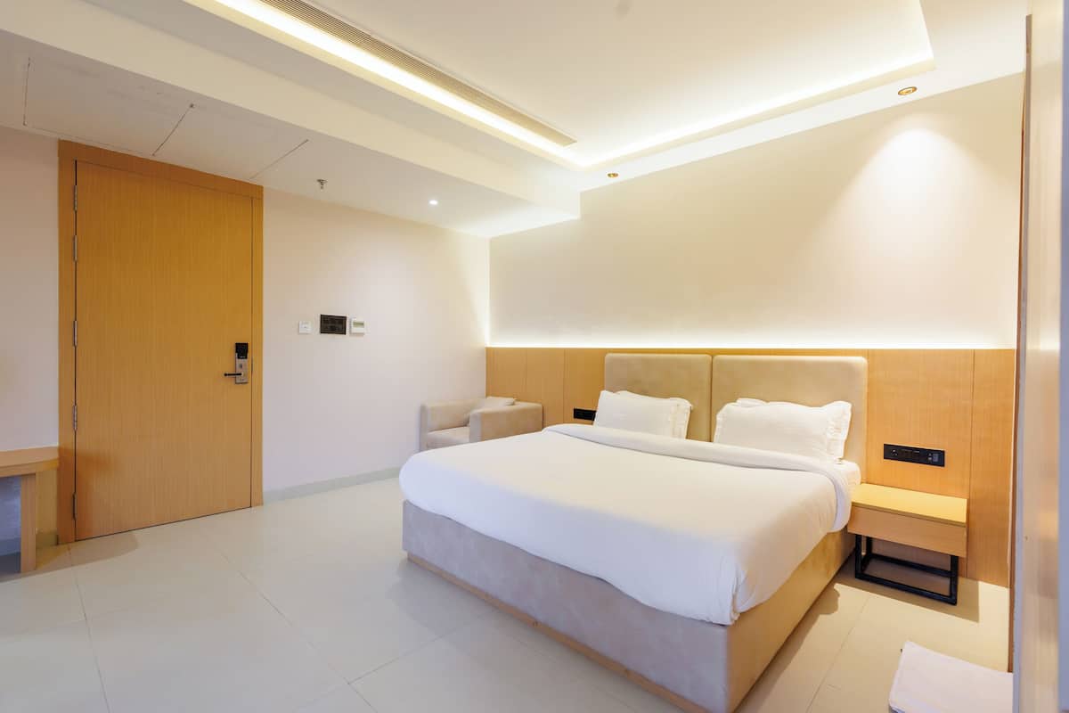 A modern hotel room with a large bed, bedside tables, a wooden wardrobe, and a small seating area.