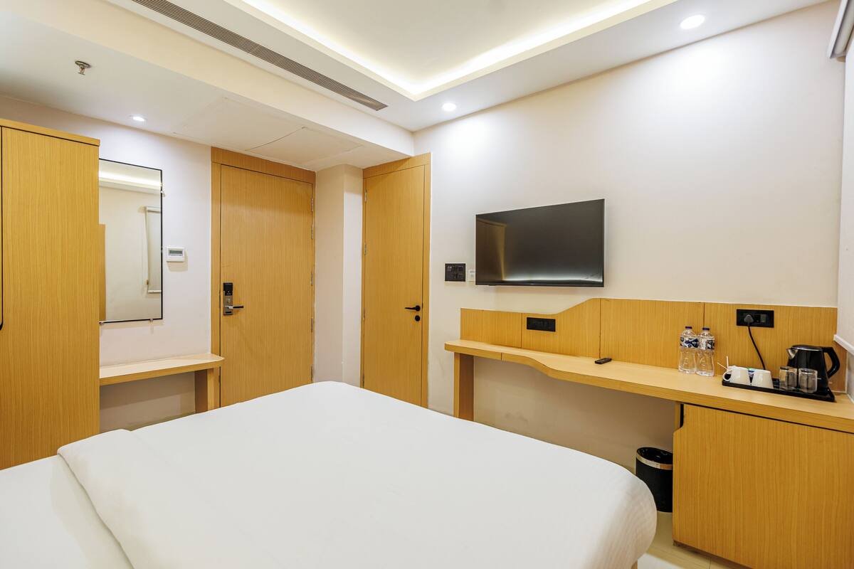 A hotel room with a bed, a desk with a flat-screen TV, a mirror, and a trash can.