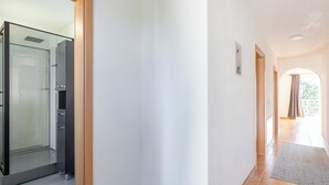 A hallway with a glass door leading to a shower area, a tiled floor, and a view of a garden through an open door. - 90 sqm Cozy home cinema with balcony & projector (Kassel)